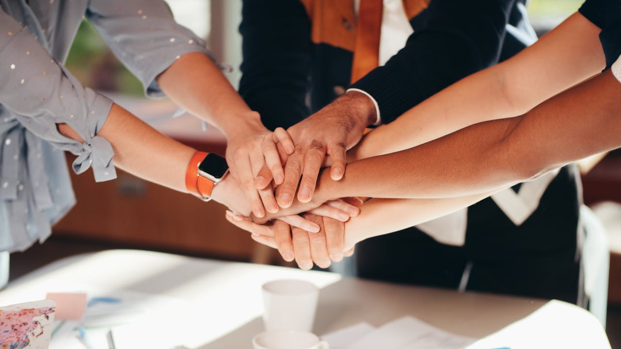 Services A close-up shot of diverse team members stacking hands over a white table, symbolizing unity and collaboration.