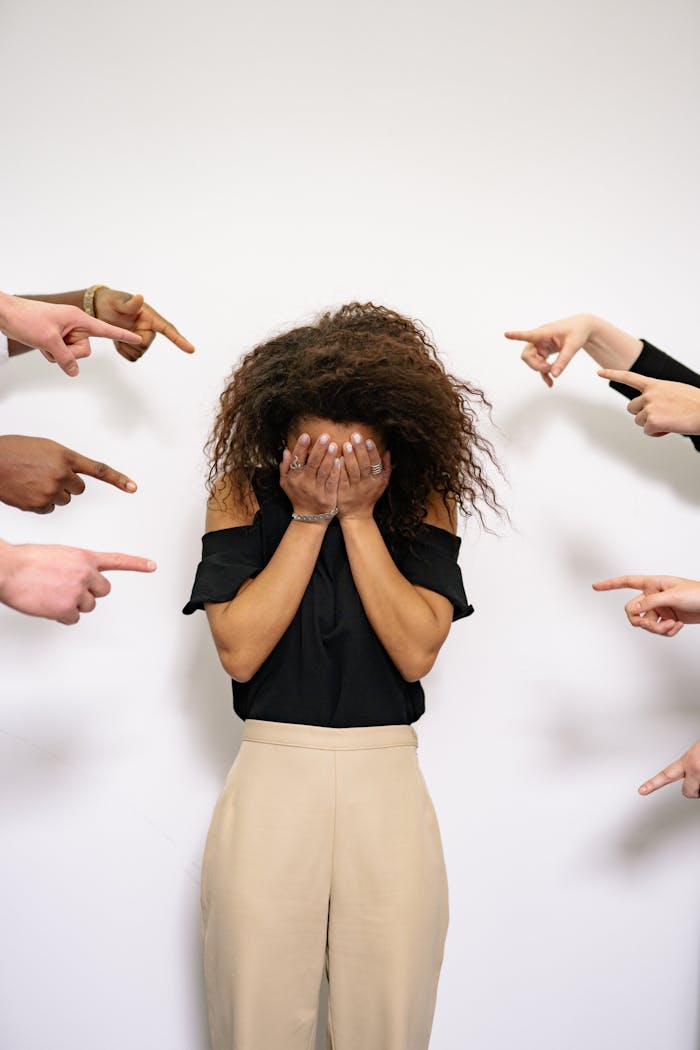 Home A stressed woman covers her face as multiple hands point accusatorily in a studio setting.